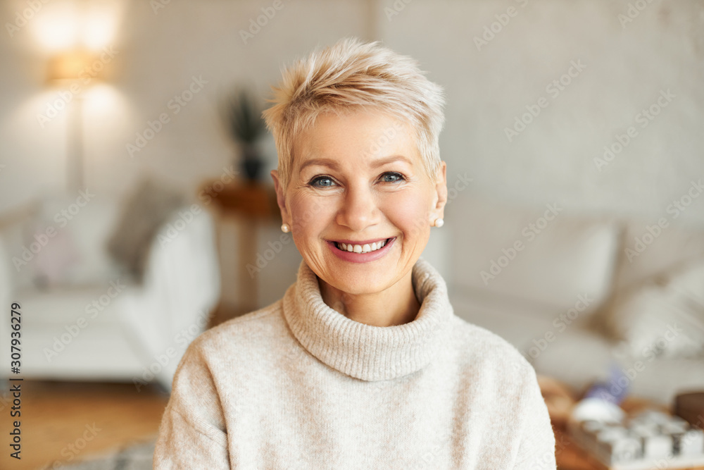 http://Close%20up%20image%20of%20happy%20good%20looking%20elegant%20fifty%20year%20old%20woman%20wearing%20warm%20cozy%20jumper,%20pearl%20earrings%20and%20short%20stylish%20hairdo%20being%20in%20good%20mood%20sitting%20in%20living%20room,%20smiling%20broadly%20at%20camera